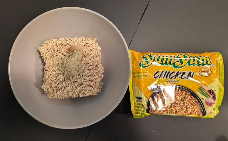 Trockener Nudelblock auf einem grauen Teller, darauf das Würzpulver, daneben die Yum Yum Chicken Packung